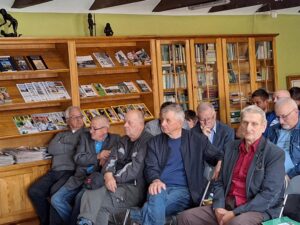 Spotkanie historyczne, grupa ludzi słucha prelekcji. Widać ścianę z książkami.