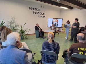 Spotkanie historyczne, grupa ludzi słucha prelekcji. Widać ekran multimedialny oraz ścianę z napisem "80-lecie polskich Lipian"