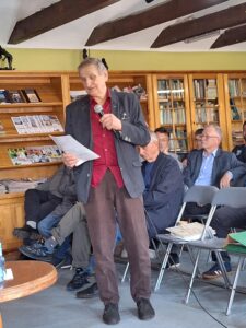 Spotkanie historyczne, grupa ludzi słucha prelekcji. Widać ścianę z książkami.